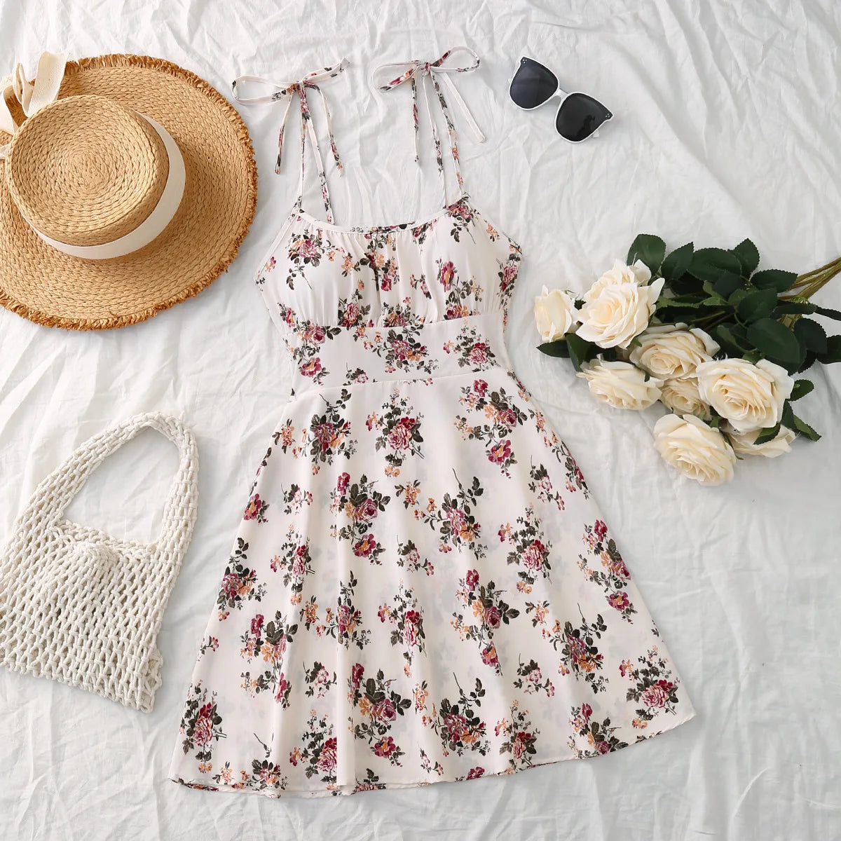 Summer French style white mini dress with pink floral print, tie straps, and elastic waist on a flat lay background with holiday accessories.